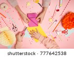 Colorful pink background with various party confetti, paper decoration, flags, stationary, DIY accessories with woman's and kid's hands making greeting card. Fat lay top view. Party arrangement