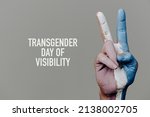 the text transgender day of visibility and the hand of a person doing the V-sign with the transgender pride flag painted in it, on a pale gray background
