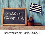 Small photo of a chalkboard with the question question do you speak greek? written in greek, a pot with pencils, an eraser and the flag of Greece, on a wooden desk