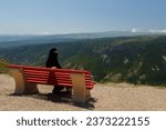 A Muslim woman in a black niqab sits on a bench and looks at the mountains from the back. Horizontal photo.