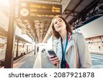 Small photo of Young woman with departure times behind her waiting for her train while holding her mobile phone - Woman looking at the clock in the train station while her train is delayed - Transportation concept