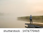 Young man standing alone on wooden footbridge and staring at lake. Mist over water. Foggy air. Early chilly morning. Empty place for sentimental, inspirational text, quote or sayings. Back view.
