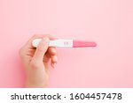 Young woman hand holding pregnancy test with two stripes. Pastel pink color background. Positive result. Closeup. Point of view shot. 