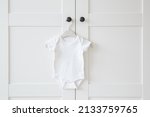 White baby bodysuit hanging on hanger at door of wardrobe. Closeup. Front view. Clothes preparing for newborn.