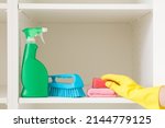 Detergent bottle, brush, rags and sponge on white shelves inside opened wardrobe. Woman hand in rubber protective glove taking cleaning things. Closeup. Front view.