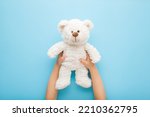 Baby boy hands holding smiling white teddy bear on light blue table background. Pastel color. Closeup. Point of view shot. Kids best friend. 