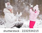 Happy little daughter and young adult mother throwing fresh white snow at park. Lovely fun moment. Spending time together and enjoying cold winter day. Side view.