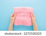 Young adult woman hands holding pink folded dry soft microfiber rag for different surfaces wiping. Closeup. Light blue table background. Pastel color. Point of view shot.