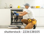 Repairman in uniform repairs dishwasher in kitchen. Young man specialist unscrews parts with screwdriver checking state of trays for dish side view.