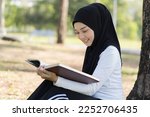 Asian young Muslim woman wear hijab black headscarf and hijab dress sitting and reading a book in the park