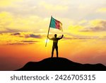 Small photo of Cameroon flag being waved by a man celebrating success at the top of a mountain against sunset or sunrise. Cameroon flag for Independence Day.