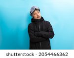 Small photo of Winter sport. Handsome asian man with snowboard goggles and black anorak coat with curious face stand on blue background.