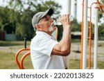 Small photo of Healthy mature man in his 60th in sports t-shirt drinking bottled water to restore water balance after workout on iron outdoor simulator on special sports ground, oldster keeping healthy lifestyle