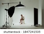Young barefoot man sits on chair in pose of thinker in photo studio. Male model wearing white shirt on white background among studio equipment and lighting fixtures. Backstage concept.