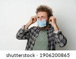 Small photo of Disgustedly under duress puts on protective medical mask on his face to protect coronavirus, with curly hair, young man wearing plaid shirt and olive t-shirt under. White background.