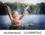 little girl smiling playing in the river. A girl with blond hair raises her hands up in the water and splashes water drops.