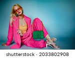Fashionable woman wearing trendy summer pink fuchsia color suit, strappy sandals, orange sunglasses, with green quilted faux leather bag, posing in studio, on blue background. Copy, empty space