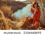 Happy smiling fashionable woman wearing trendy orange color hat, autumn trench coat, brown high boots, with white faux leather bag, posing in nature. Copy, empty space for text