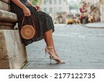 Trendy summer rotan wicker bag, white strap sandals in stylish female outfit. Woman posing in street of European city. Fashion details. Copy, empty space for text