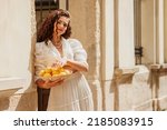Beautiful curly brunette woman wearing trendy summer white linen shirt, skirt, holding bowl with lemons, posing in sunny street of European city. Copy, empty space for text