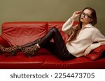 Small photo of Fashionable woman wearing trendy zip neck knitted sweater, glasses, brown leather leggins, ankle boots with leopard print, posing, laying on couch. Studio fashion portrait. Copy, empty space for text