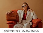 Small photo of Fashionable confident woman wearing elegant white suit, sunglasses, chunky chain, holding classic brown leather bag, sitting in armchair, posing on beige background. Copy, empty space for text