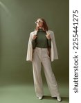 Small photo of Fashionable confident woman wearing elegant white suit with blazer, wide leg trousers, cashmere turtleneck sweater, trendy sunglasses, posing on green background. Full-length studio fashion portrait