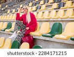 Fashionable confident blonde woman wearing trendy pink sunglasses, fuchsia color coat,   zebra print trousers, white sneakers, holding faux leather tote, shopper bag, posing outdoors, on stadium seat