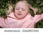 Small photo of Portrait of caucasian blonde girl with closed eyes on green grass in pink jacket . Towheaded little girl outside