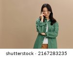 Serious adorable Korean young woman in khaki green shirt stylish eyewear looks at camera posing isolated on over beige pastel studio background. Cool fashion offer. Sunglasses ad concept