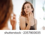 Small photo of Portrait of a young woman touching a pimple on her face while looking in the mirror. Facial skin problems, medical care and treatment concept.