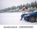 A smiling woman drinks a hot drink from a thermos while standing near her car on a winter snowy road in the forest. The concept of rest, freedom, relaxation, travel.