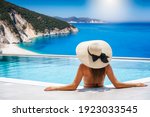 A woman in bikini and with hat sits in the swimming pool and enjoys the view to the turquoise sea of Kefalonia island, Ionian Sea, Greece, during summer holiday time