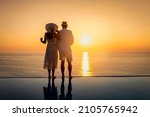 A hugging couple stands by the swimming pool and enjoys the intense sunset over the mediterranean sea in Greece with a glass of wine