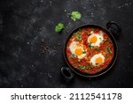 Small photo of Shakshuka eggs in a pan with toast on a black concrete background. Poached eggs in a spicy tomato pepper sauce. Traditional Jewish scrambled eggs. Top view, flat lay. Textured object, selective focus