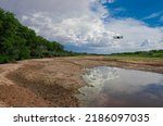 Small photo of Dried up river bed with drone flying over top with clouds and blue sky in the distance and green trees along the edge of the dried up Rio Grande