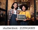 Two young startup barista partners with aprons stand at casual cafe door, letters on board and show open sign, happy and cheerful smiles with coffee shop service jobs, and new business entrepreneurs.