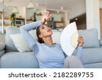 Small photo of Woman puts head on sofa cushions feels sluggish due unbearable heat, waves hand fan cool herself, hot summer flat without air-conditioner climate control system concept