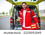 Small photo of Young man , a paramedic, standing at the rear of an ambulance, by the open doors. He is looking at the camera with a confident expression, smiling, carrying a medical trauma bag on his shoulder.