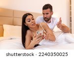 Small photo of Photo of young couple in bed, woman use smartphone try explain envy angry husband she dont have another man point hand screen feel confused. Jealous husband spying his wife mobile phone