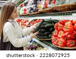 Small photo of Woman buy vegetables in grocery store, supermarket. Caucasian lady holding marrow vegetable and choosing from open refrigerator.