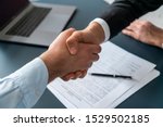 Close up of two businessmen shaking hands in blurred office with computer and contract on table. Concept of partnership and communication