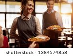 Two waiters serving lunch and brining food to their gusts in a tavern. Focus is on happy waitress. 