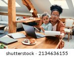 Small photo of African American little girl using cell phone and sitting on mother's lap while she going through paperwork and working on laptop at home.