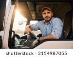 Small photo of Young truck driver sitting behind steering wheel in a cabin and looking at camera.