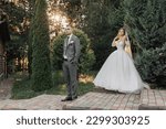 Small photo of portrait of the bride and groom in nature. The groom in a gray suit is in the foreground, the bride is walking behind him, smiling in a white voluminous dress, holding a bouquet. Stylish groom