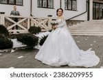 Small photo of The bride in a white voluminous dress with a long train, holds a bouquet in her hands and poses against the background of the building. Winter wedding.