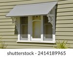 Small photo of PERIOD STYLE FEATURE AWNING WINDOW WITH DECORATIVE FRETWORK TRIMS - A timber framed window and ornate detailed shade awning with gray metal roofing on an olive green colored siding weatherboard home