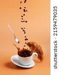 Small photo of White coffee cup, plate and metal tea spoon falling on beige table background and coffee splashes because of hit, coffee beans levitate, fly and fall to cup, crispy fresh croissant. Breakfast concept