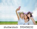 Three young beautiful girls in white dresses and wreaths of wild flowers doing selfie standing in a field. Summer in the village. Free space